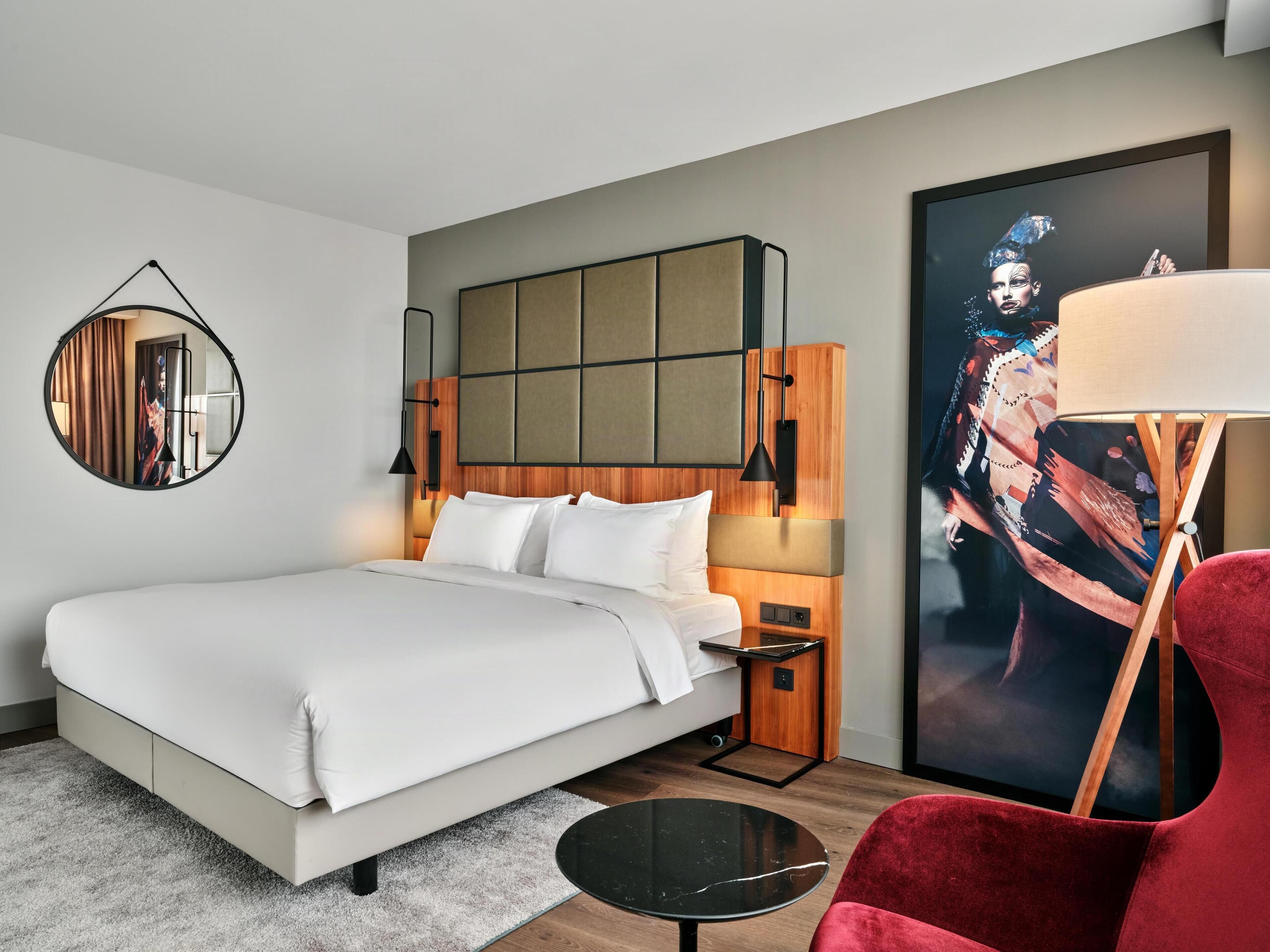 Ein modernes Hotelzimmer mit einem großen Bett, einem runden Spiegel, einem Nachttisch, einem roten Sessel, einem kleinen Tisch und einem großen Poster an der Wand.