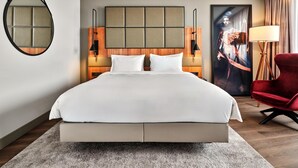A modern hotel room with a large bed, a round mirror, a wooden headboard, two wall-mounted lamps, a red chair, and a large poster on the wall. - Radisson Blu Hotel, Hamburg Airport (Hamburg)