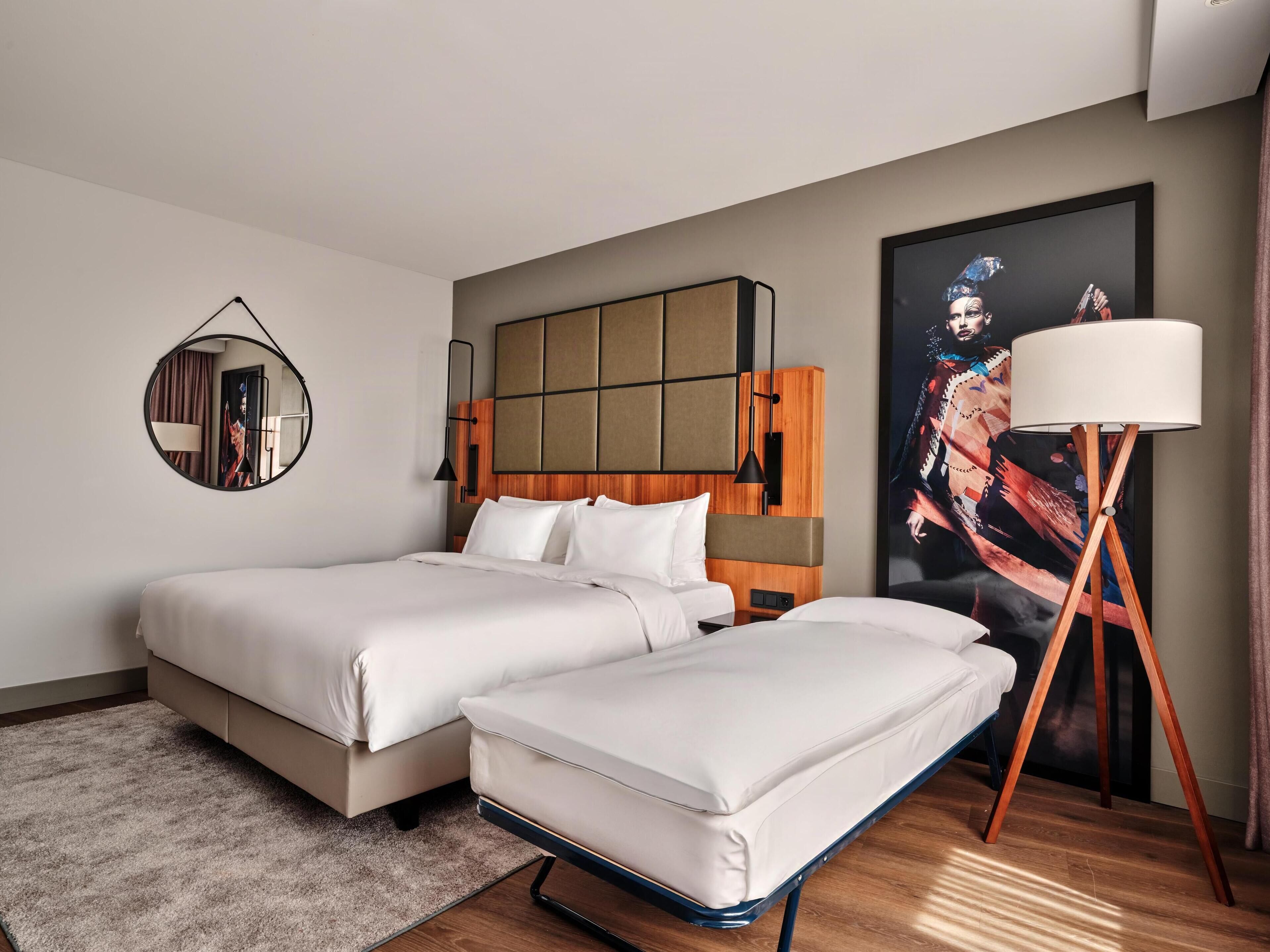 A hotel room with two beds, a large mirror, a modern lamp, and a large painting on the wall.