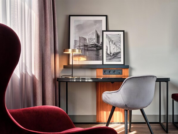 A modern hotel room with a desk, a red chair, and two framed black and white photographs on the wall.