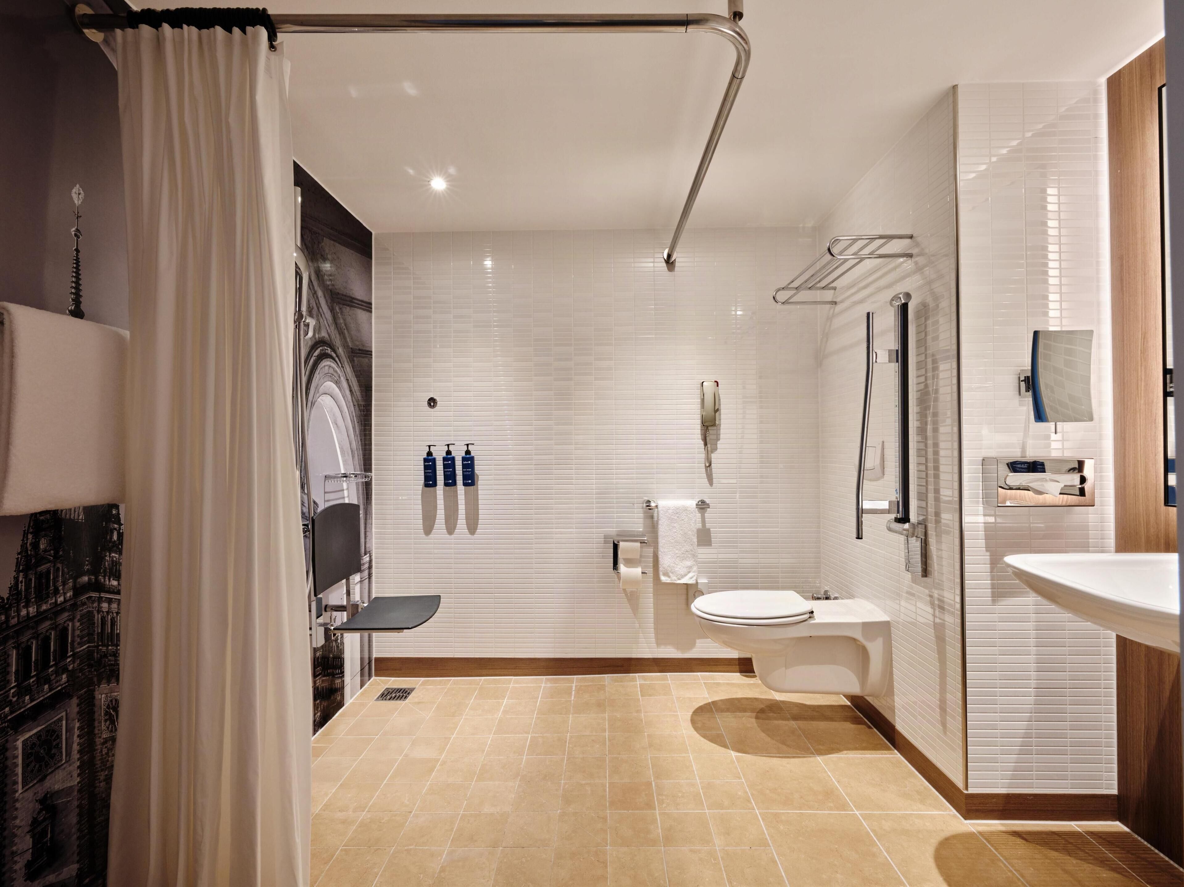 Une salle de bain moderne avec des toilettes, un lavabo et un espace douche. On y trouve trois bouteilles de savon accrochées au mur, un porte-serviettes et un miroir.