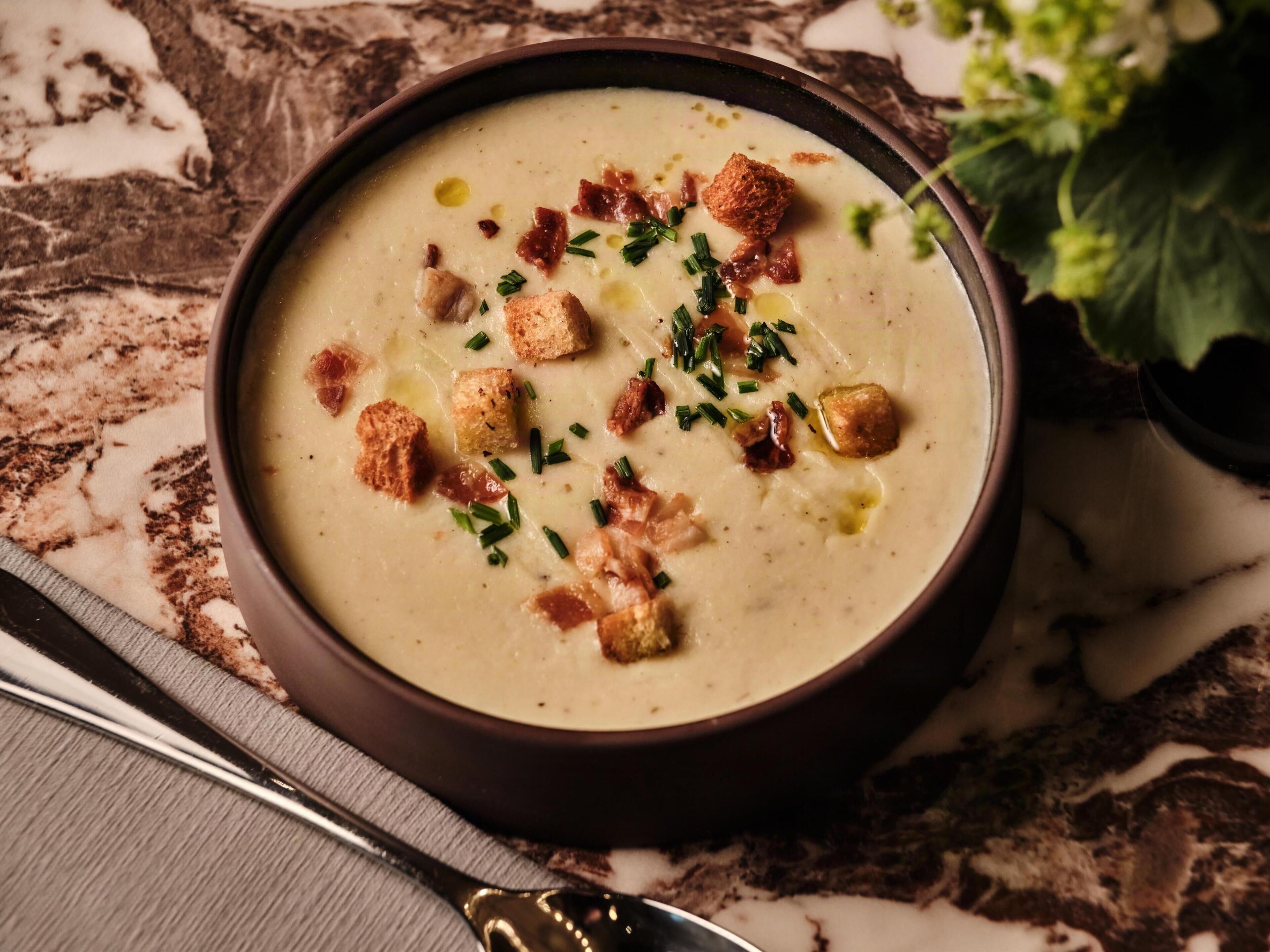 Un tazón de crema de sopa decorado con cubitos de pan y cebollín, sobre una superficie de mármol, con una cuchara a un lado.