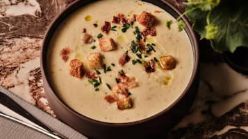 Un tazón de crema de sopa decorado con cubitos de pan y cebollín, sobre una superficie de mármol, con una cuchara a un lado.