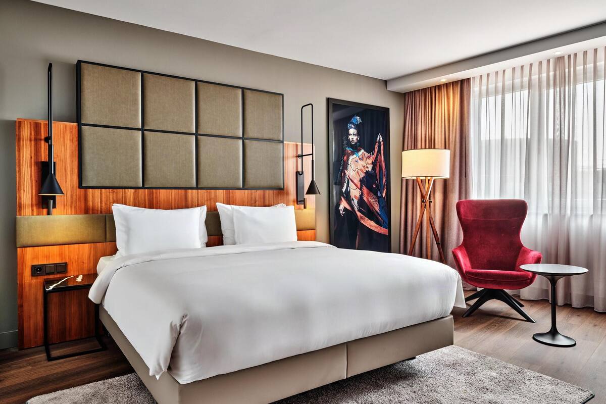A hotel room with a large bed, a red armchair, a small table, and a painting on the wall.