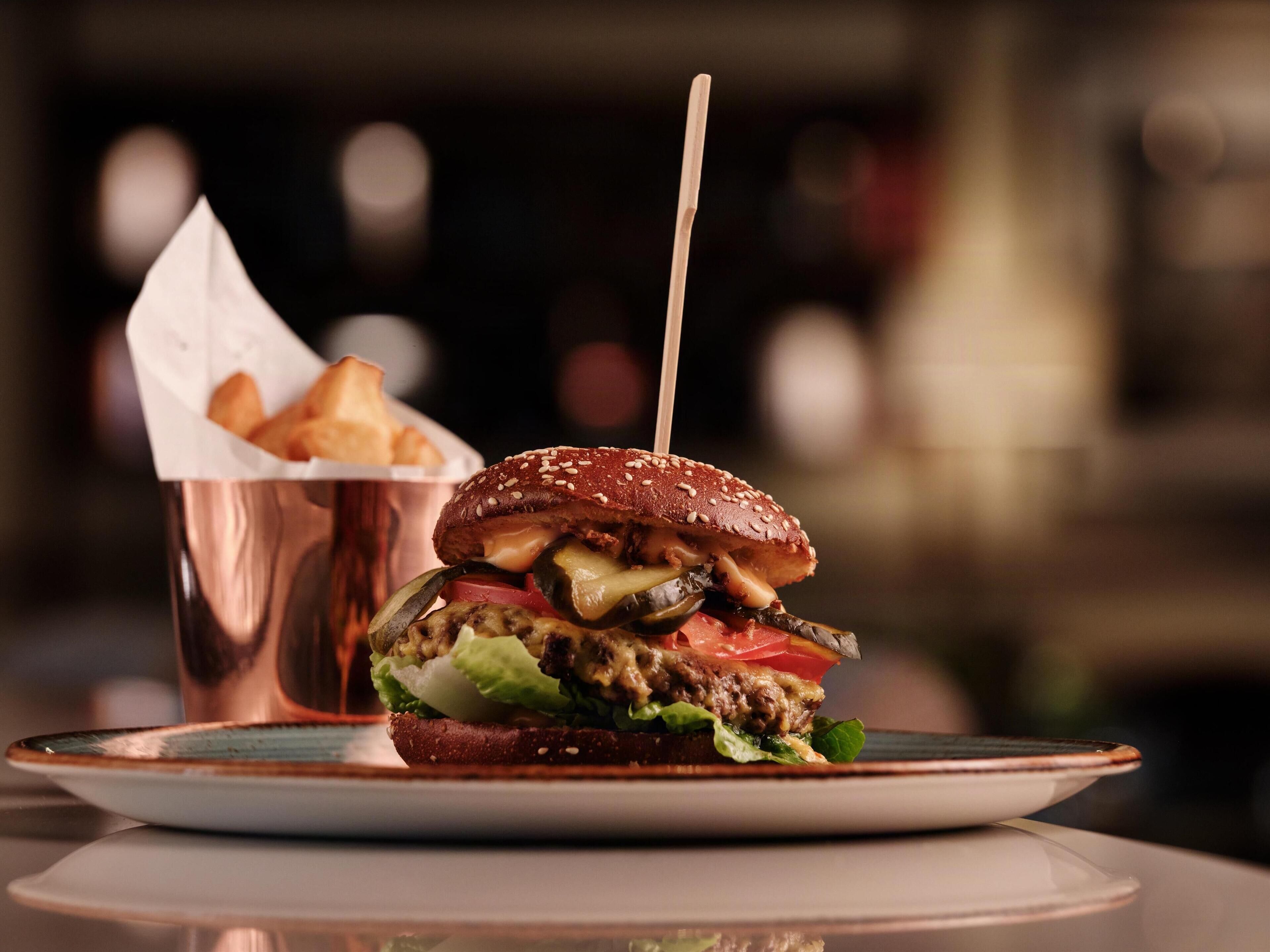 A burger with a sesame seed bun, pickles, lettuce, tomato, and cheese, served on a white plate with a copper cup of fries in the background.