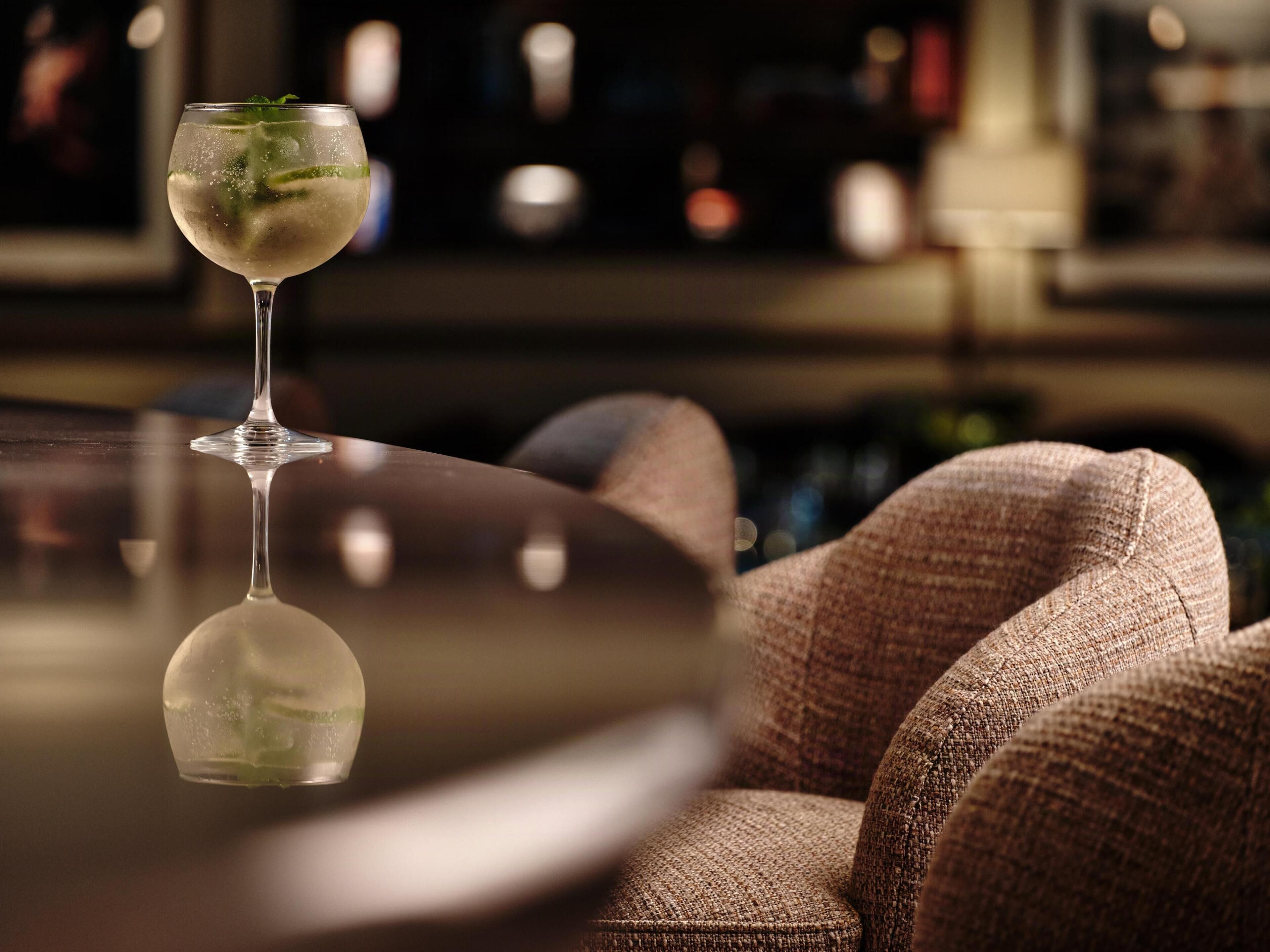 A bar area with a glass of a clear beverage, a cushioned chair, and a reflective surface.