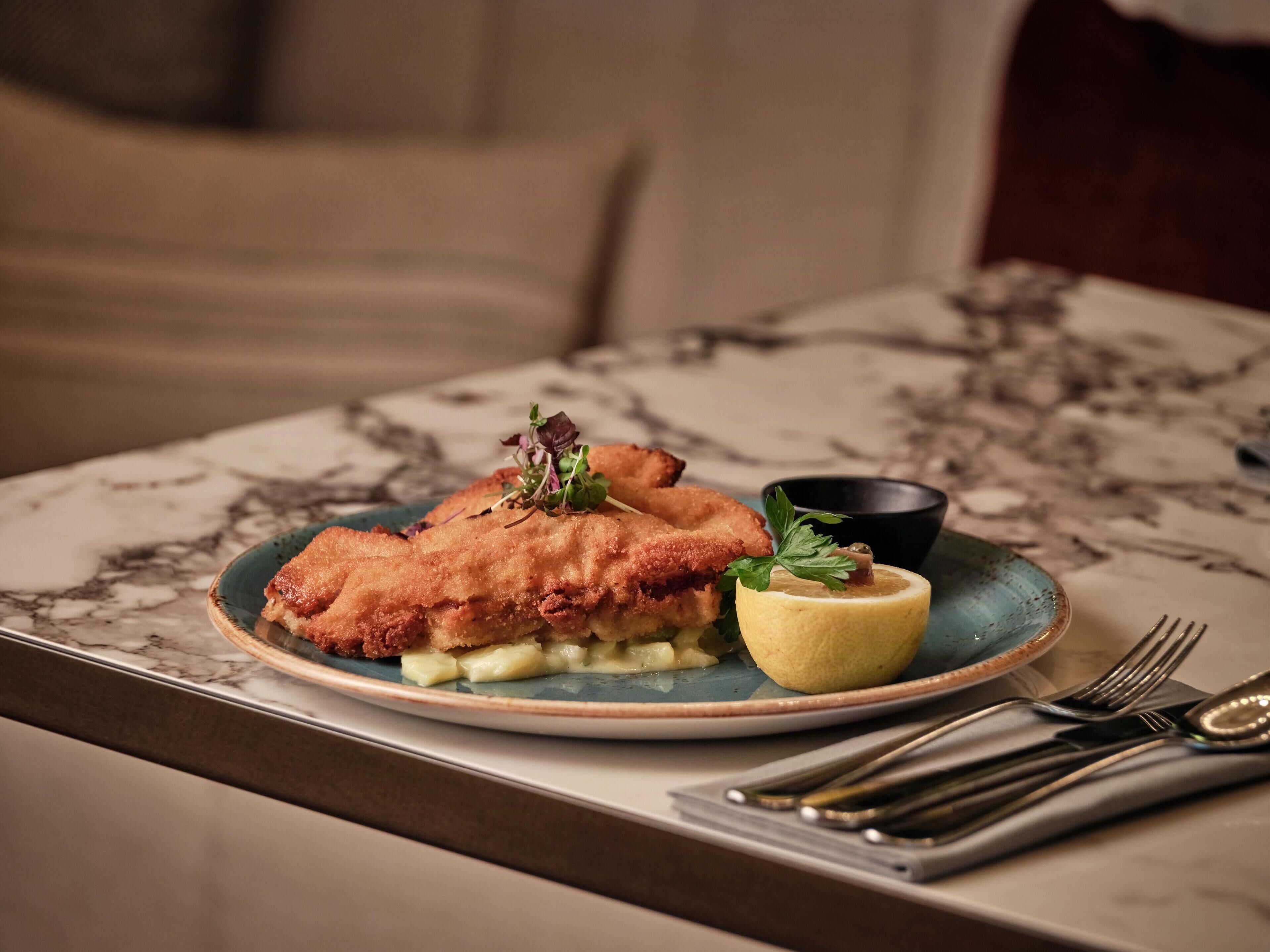 Une assiette de poisson frit, accompagnée d’une tranche de citron et d’une garniture, est posée sur une table en marbre.