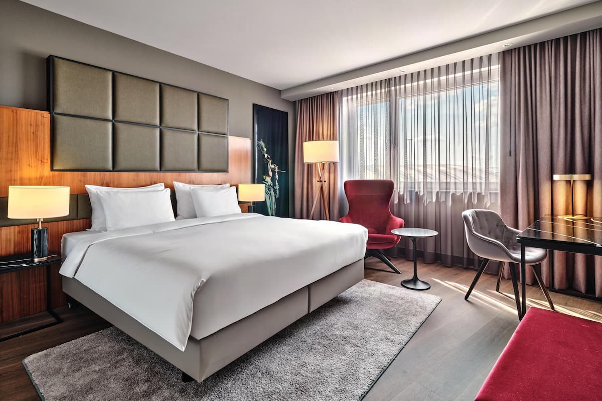 A hotel room with a large bed, a desk, two chairs, a lamp, and a view of the outdoors through the window.