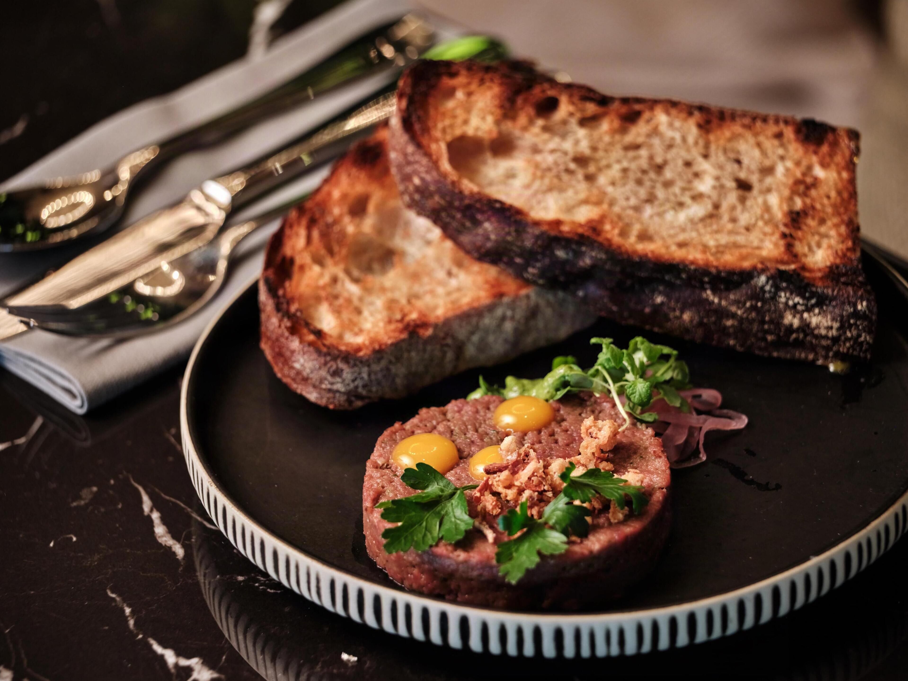 A plate with a meat dish and toasted bread, accompanied by a napkin and cutlery.