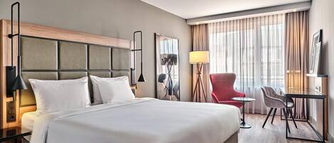 A hotel room with a large bed, a desk, a chair, a lamp, and a window with curtains.
