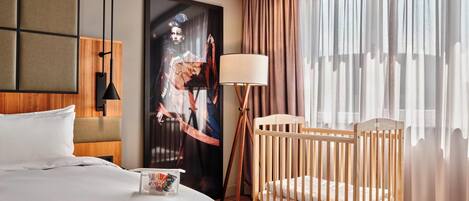 Habitación de hotel con cama, cuna, una obra de arte grande enmarcada, lámpara y vistas al exterior a través de cortinas traslúcidas.