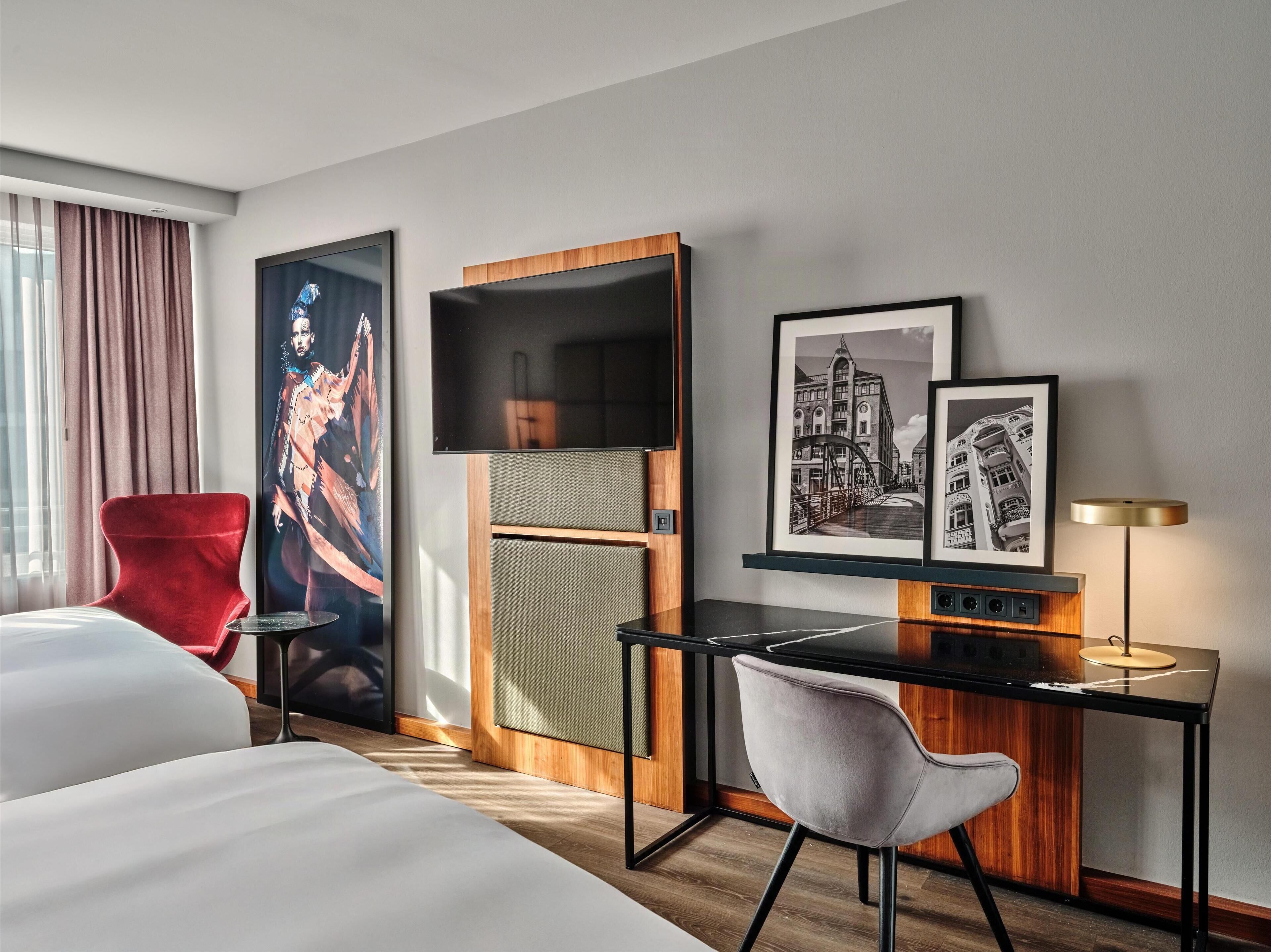 Quarto de hotel com cama, escrivaninha com cadeira, televisão e obra de arte na parede.