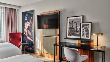 Quarto de hotel com cama, escrivaninha com cadeira, televisão e obra de arte na parede.