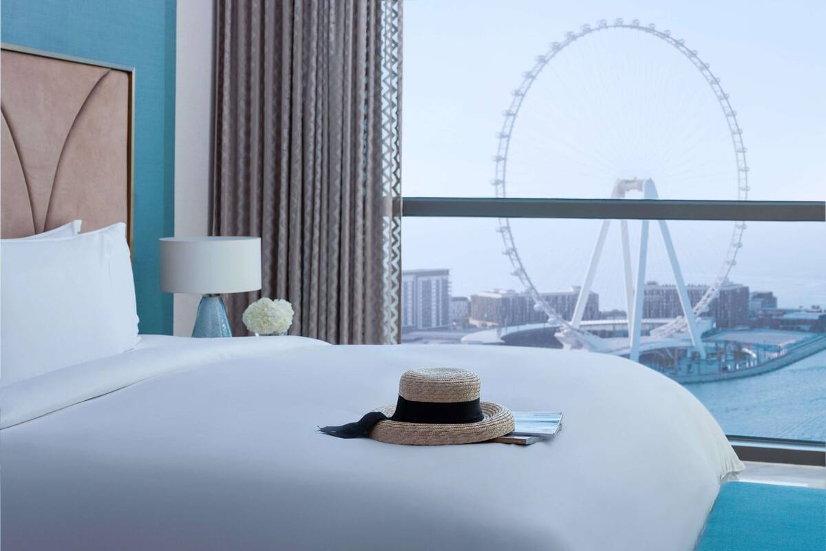 A hotel room with a bed, a lamp, a hat, and a view of a Ferris wheel.