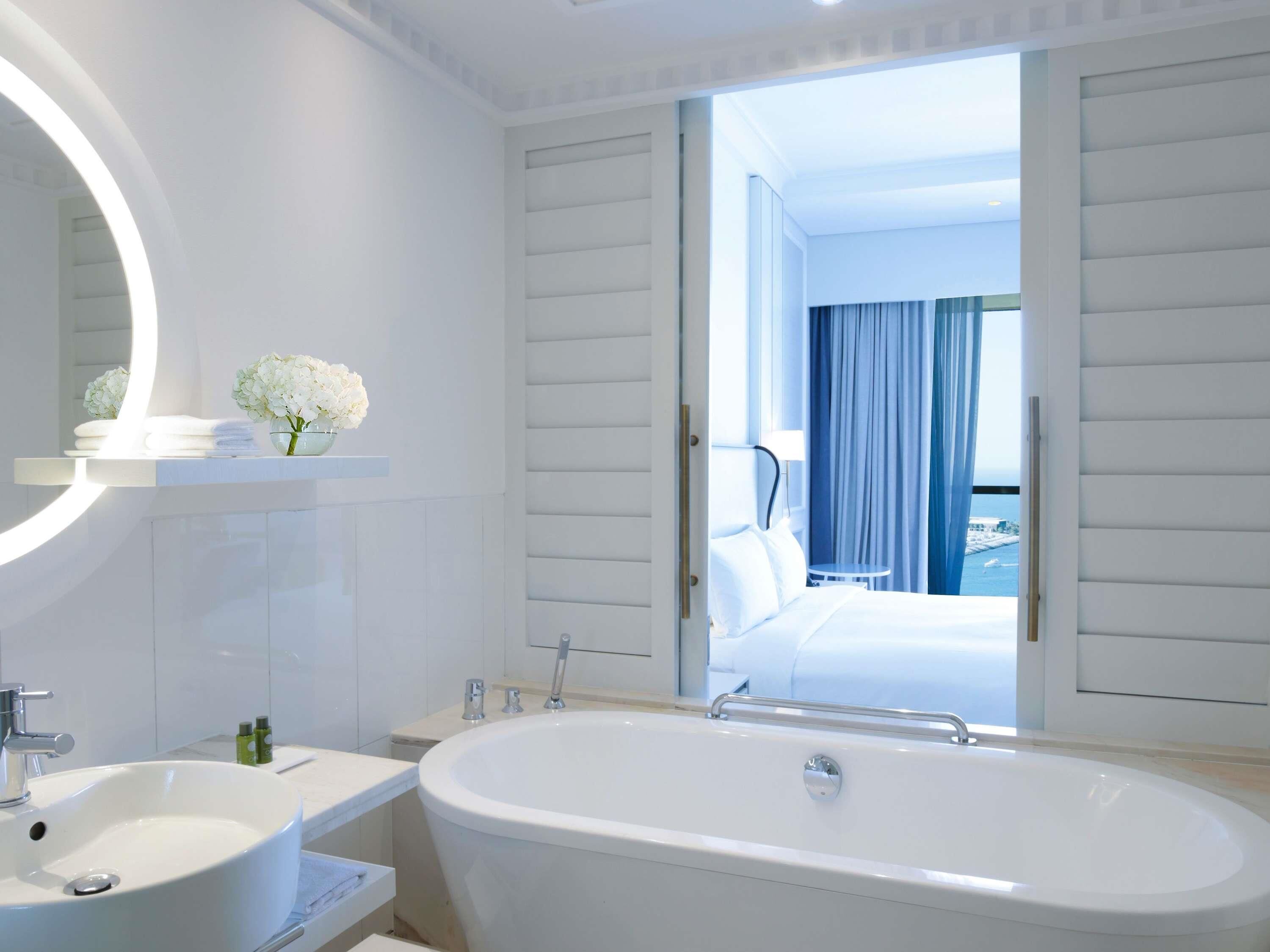 A modern bathroom with a freestanding bathtub, a large mirror, and a view of a bedroom with a bed and curtains.