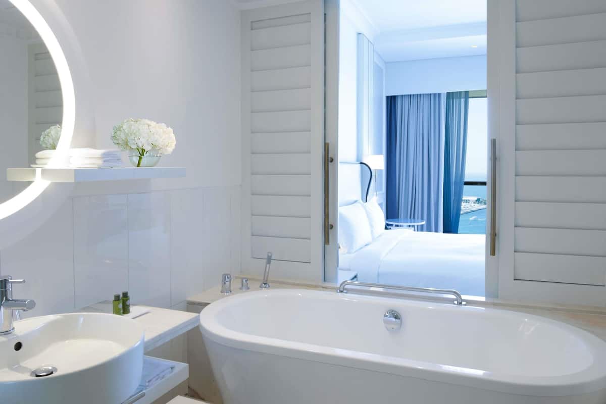 A modern bathroom with a freestanding bathtub, a large mirror, and a view of a bedroom with a bed and curtains.