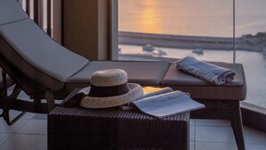 Ein Balkon mit Blick auf den Sonnenuntergang über dem Wasser, ein Liegestuhl, ein kleiner Tisch mit Hut und Handtuch sowie ein Geländer.