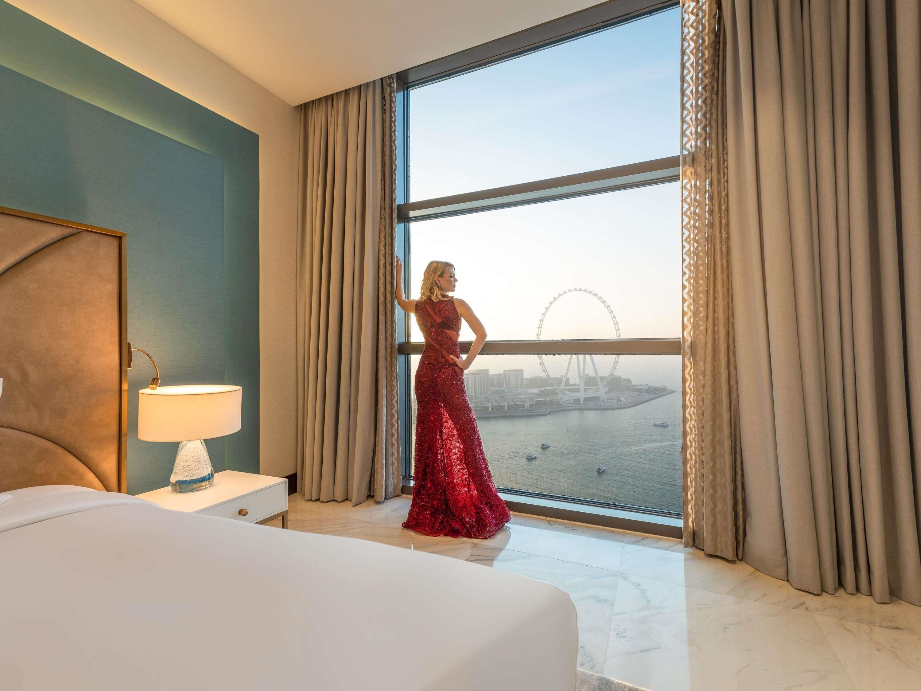 Une chambre d’hôtel avec une grande fenêtre, un lit, une table de chevet avec une lampe et une vue sur une grande roue.