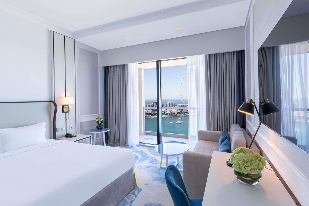 Ein Hotelzimmer mit einem großen Bett, einem Schreibtisch, einem Stuhl, einem kleinen Tisch und Blick auf Stadt und Wasser.