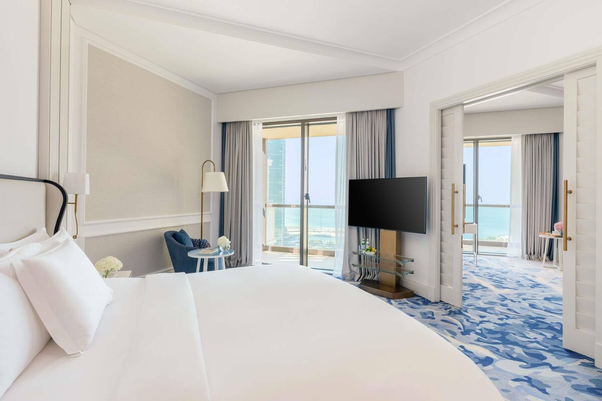 A hotel room with a large bed, a television, a small table with a lamp, and a view of the city through the balcony doors.