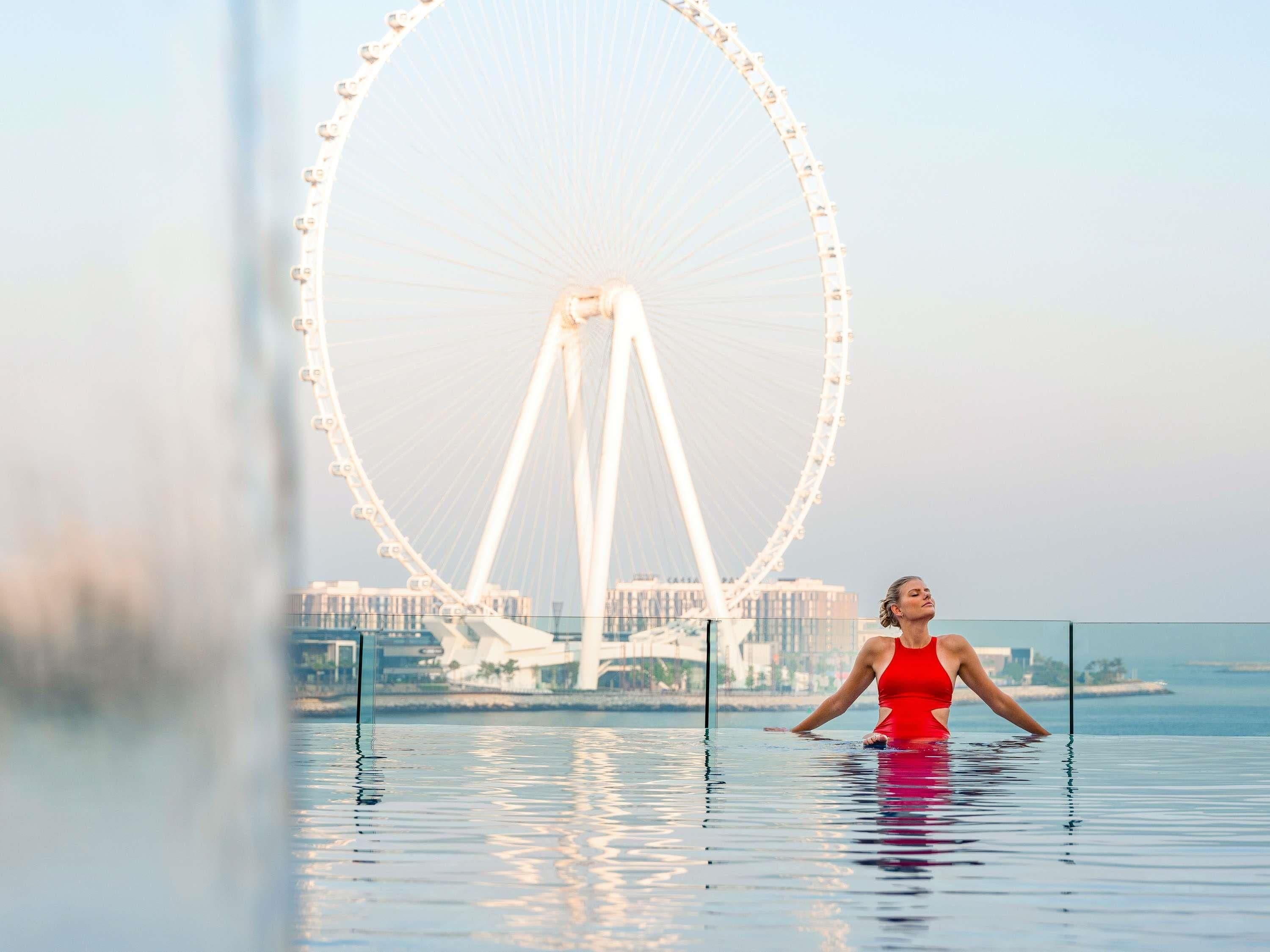 Une personne vêtue d’un maillot de bain rouge, debout dans une piscine, avec une grande roue en arrière-plan.