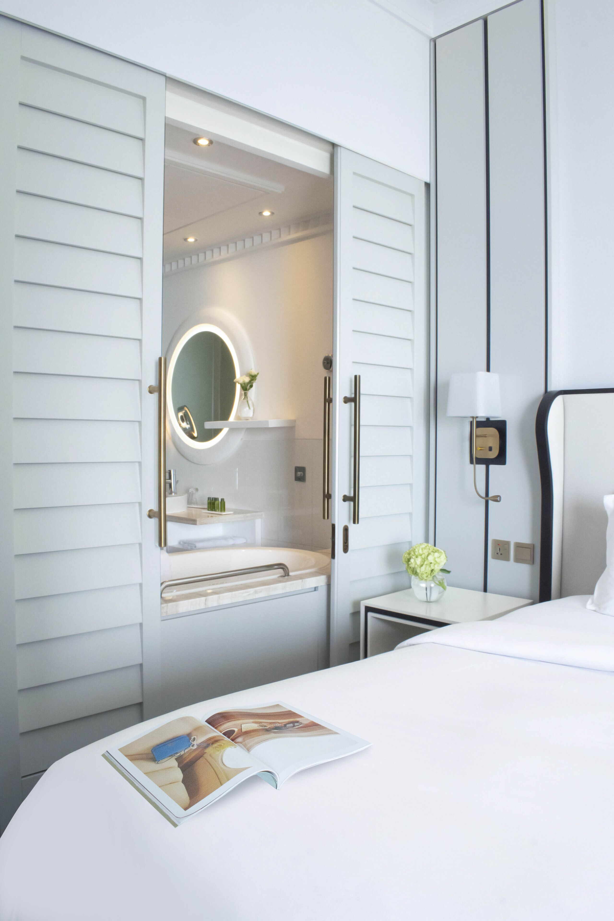 A hotel room with a large bed, a bedside table with a lamp, a vanity with a mirror, and a small table with a vase of flowers.