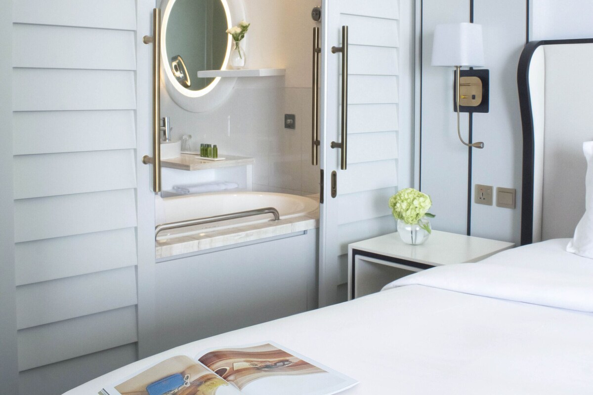 A hotel room with a large bed, a bedside table with a lamp, a vanity with a mirror, and a small table with a vase of flowers.