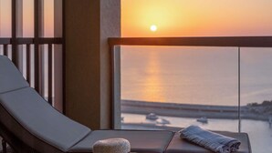 Een balkon met uitzicht op de ondergaande zon over het water, een lounge stoel en een klein rond tafeltje.