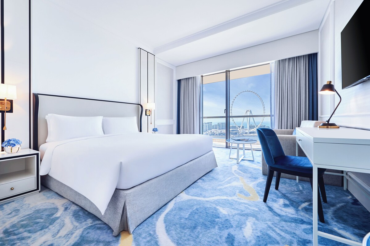A hotel room with a large bed, a desk, a chair, and a view of a Ferris wheel.