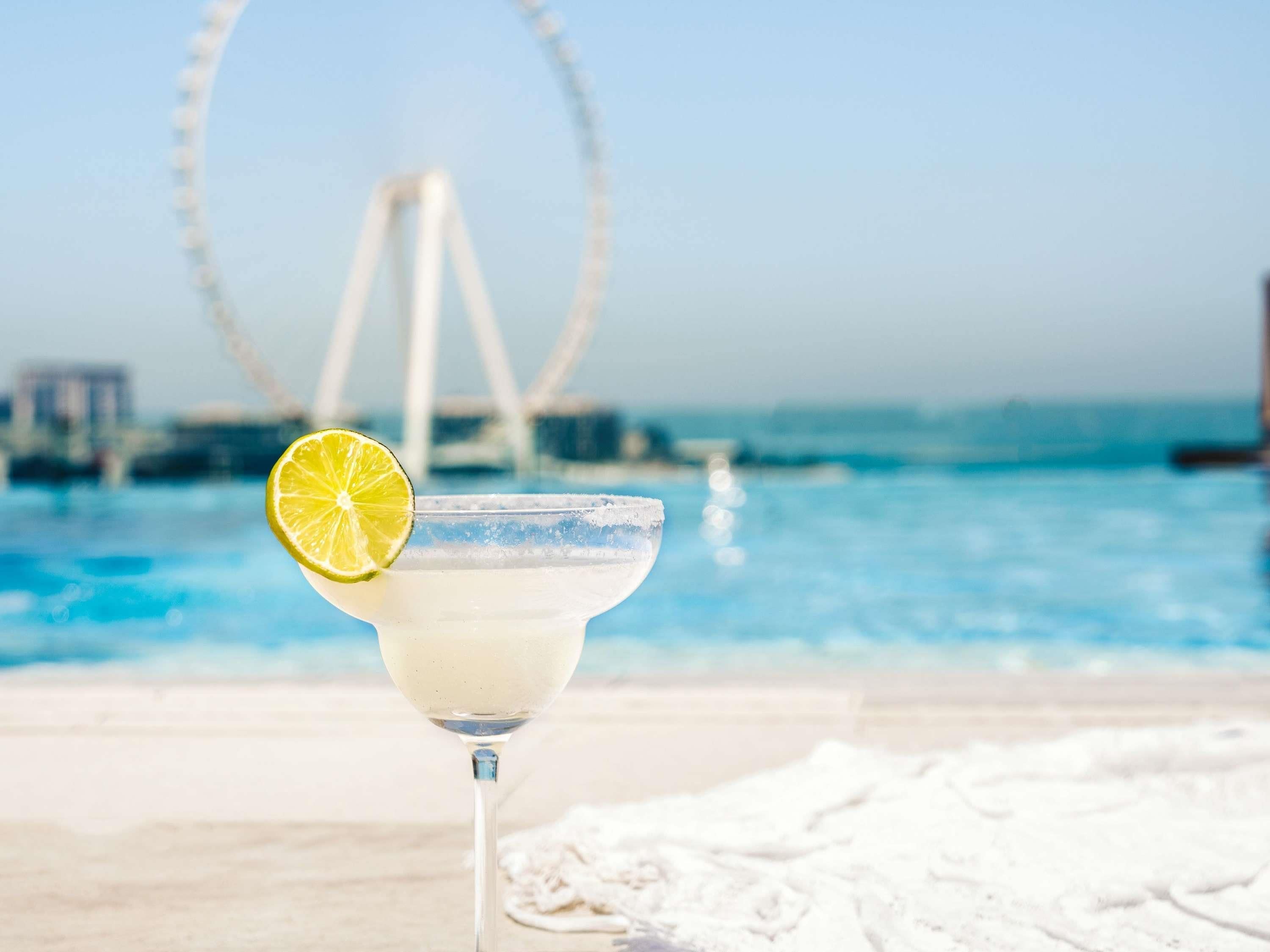 A cocktail with a lime garnish on a beach with a Ferris wheel in the background.
