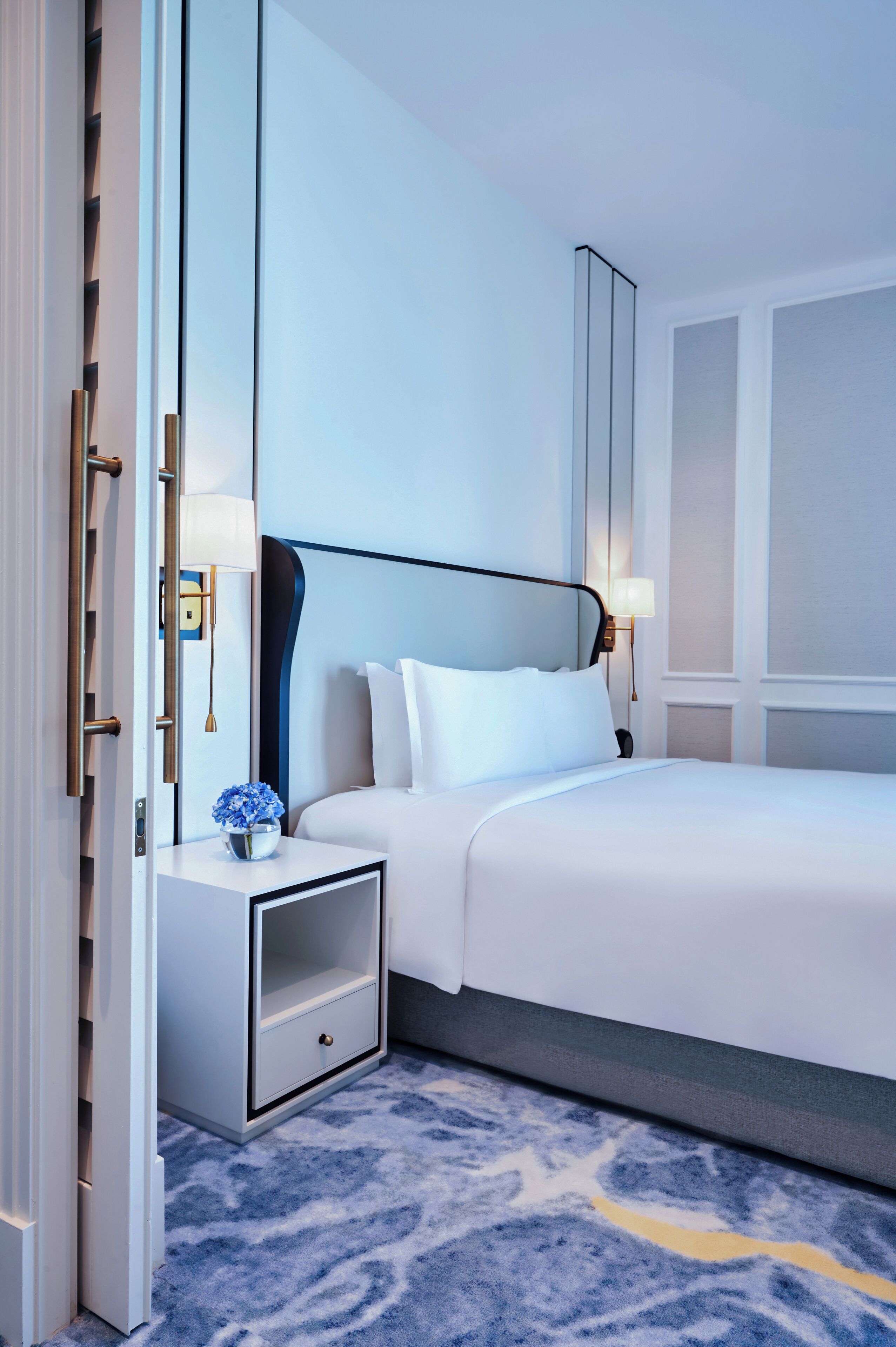 A modern hotel room with a large bed, a bedside table with a lamp, and a small side table with a vase of flowers.
