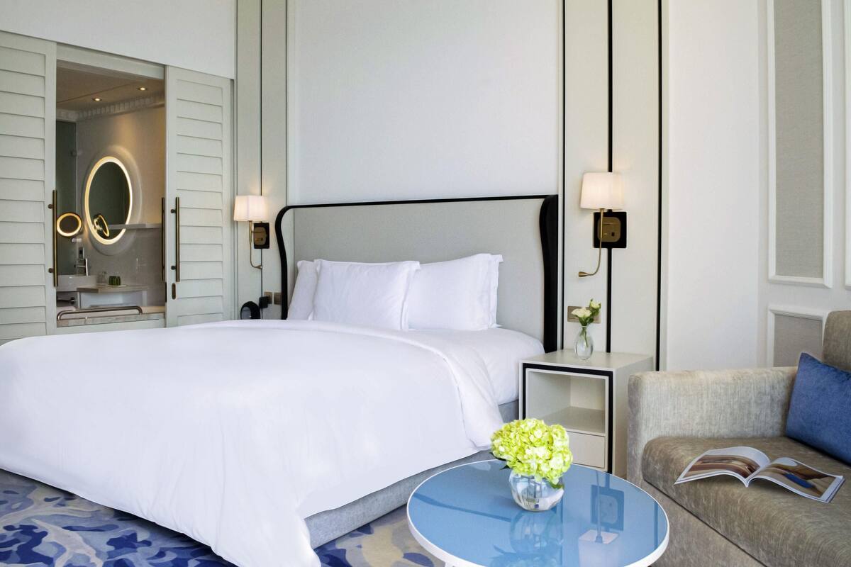 A hotel room with a bed, bedside table, lamp, and a small round table with a vase.