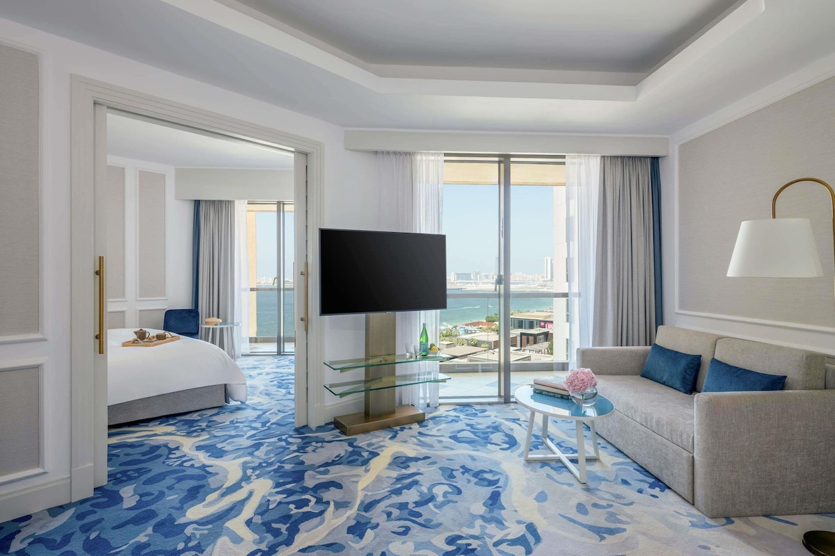 A hotel room with a bed, a TV, a sofa, a glass coffee table, and a view of the city.