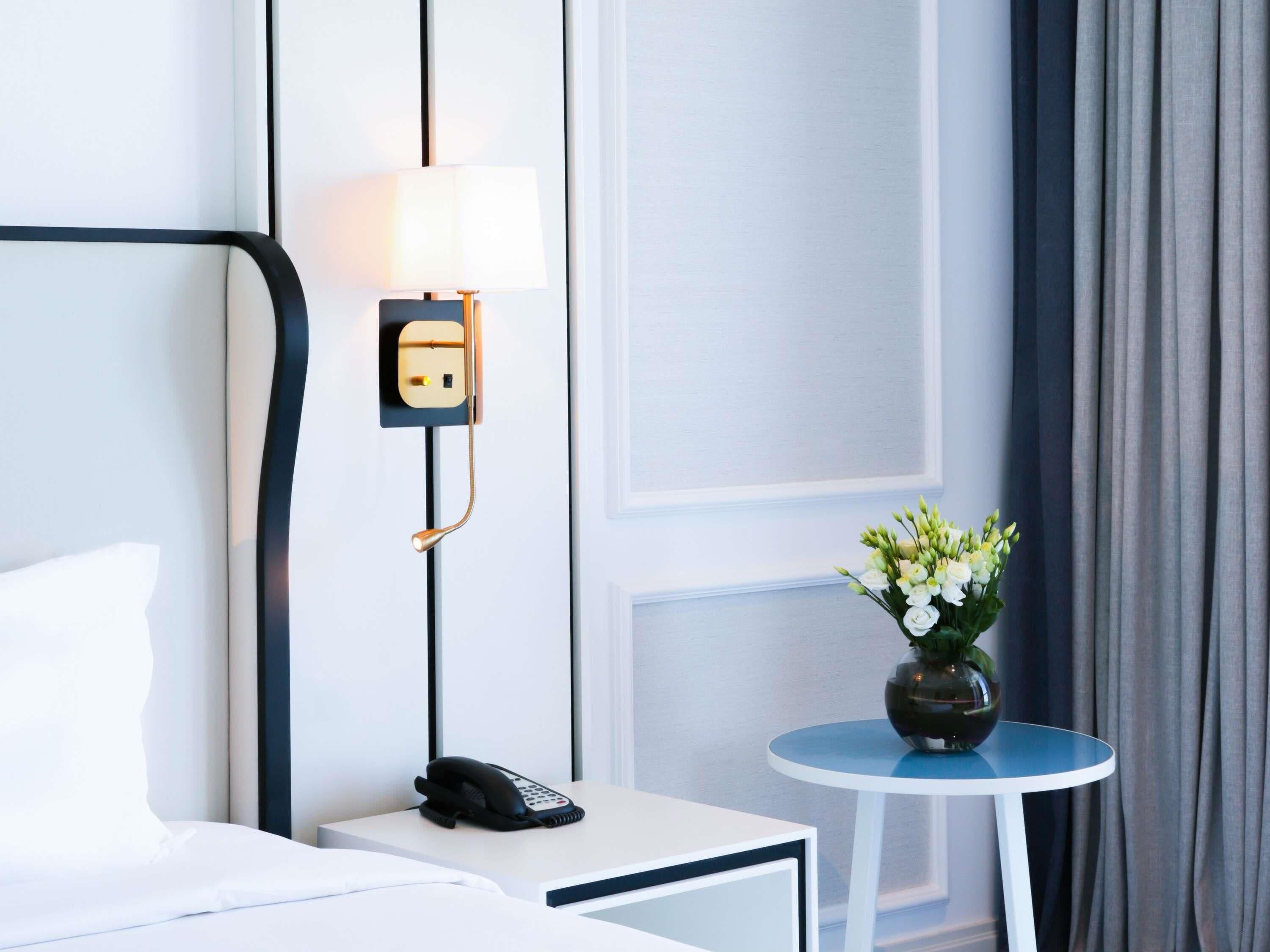 A hotel room with a bed, a bedside phone, a lamp, a small table with a vase of flowers, and a view of the outside curtains.
