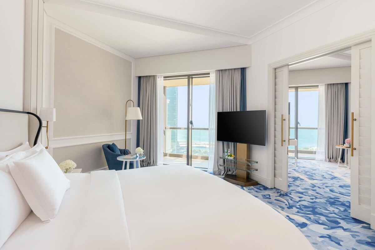 Ein Hotelzimmer mit einem großen Bett, einem Fernseher, einem kleinen Tisch mit Lampe und Blick auf die Stadt durch die Balkontüren.