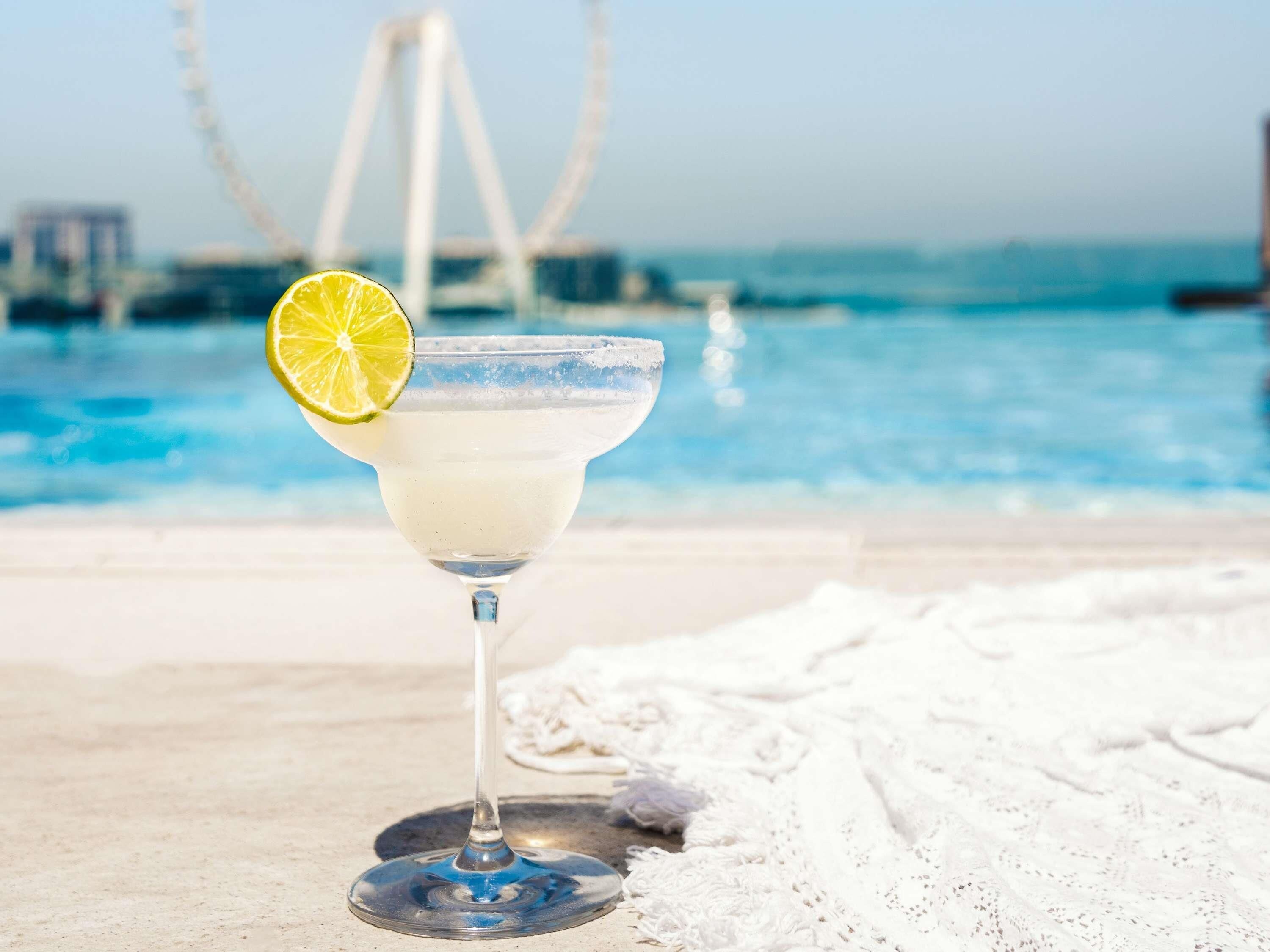 A cocktail glass with a lime garnish on a beach with a clear pool and a bridge in the background.