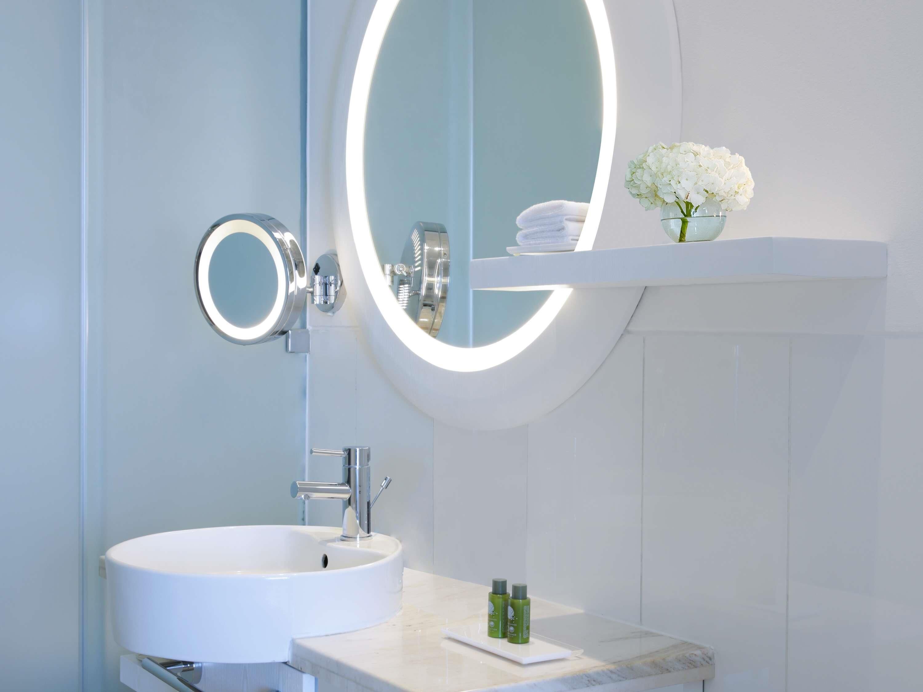 A bathroom with a white sink, a mirror with a light, and a shelf with a vase of flowers.