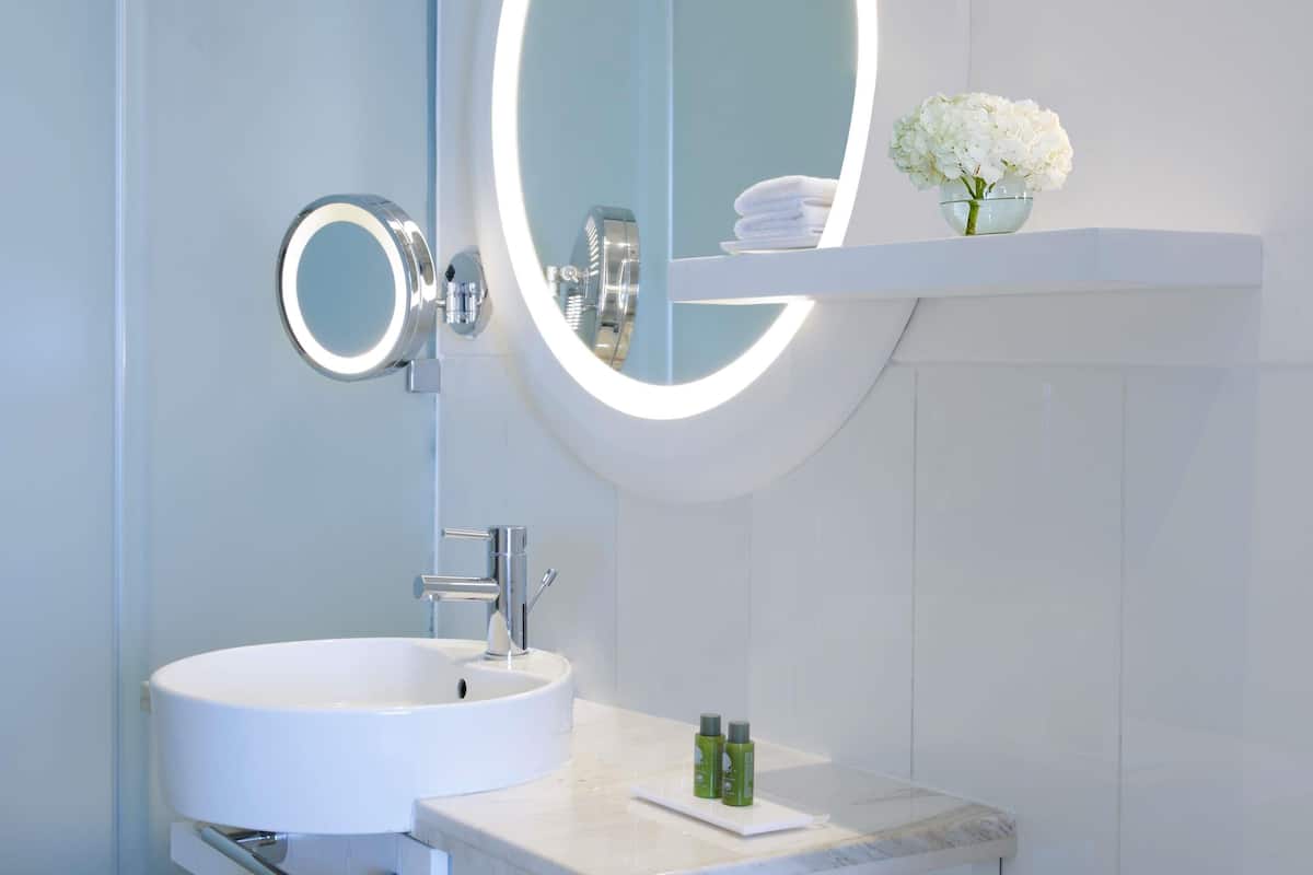 A bathroom with a white sink, a mirror with a light, and a shelf with a vase of flowers.