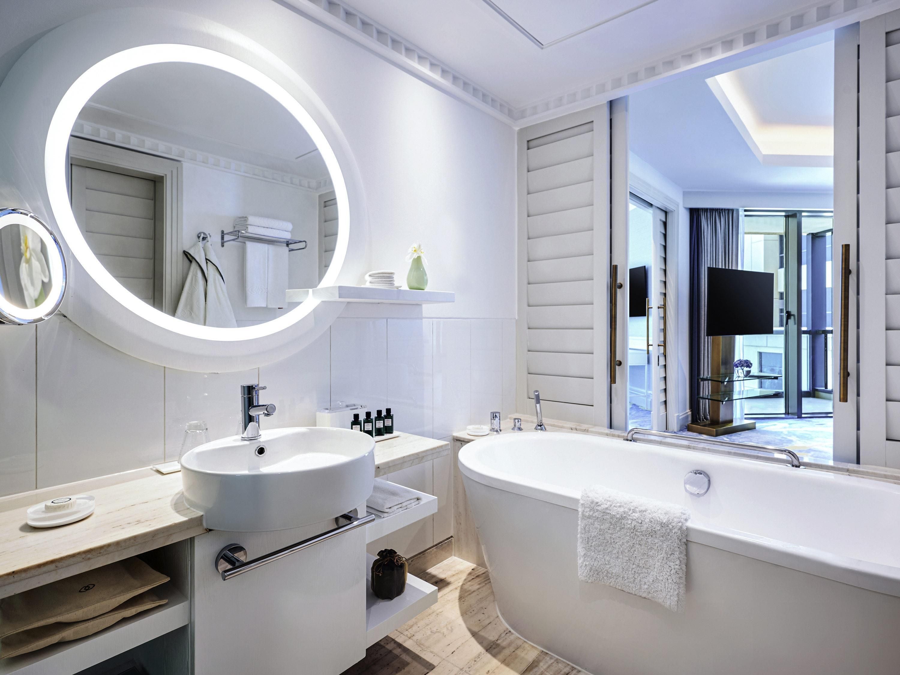 A modern bathroom with a large bathtub, a round mirror with lighting, a vanity with a sink, and a shower area.