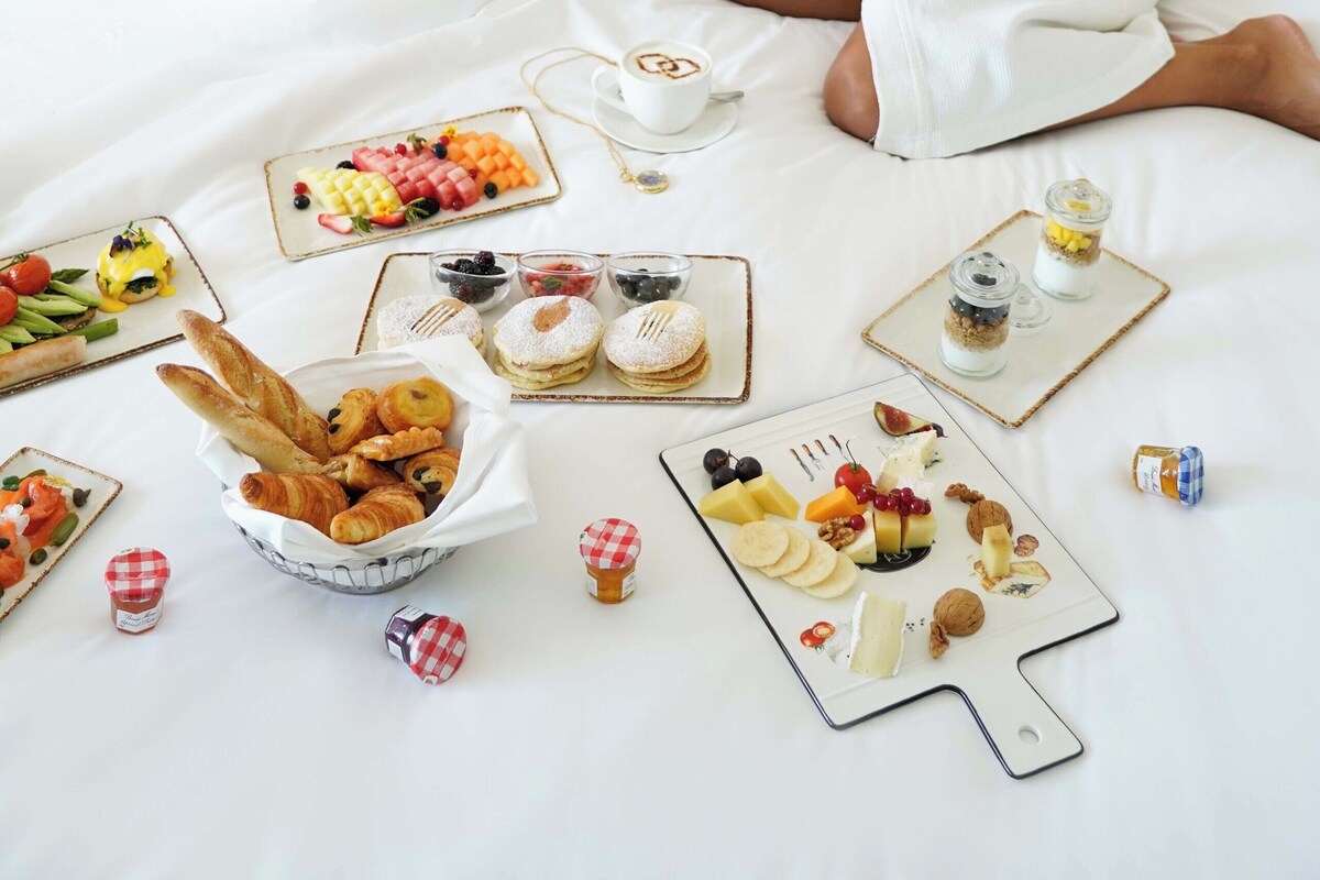Two people in white robes sitting on a bed with breakfast items like pastries, fruits, and beverages.