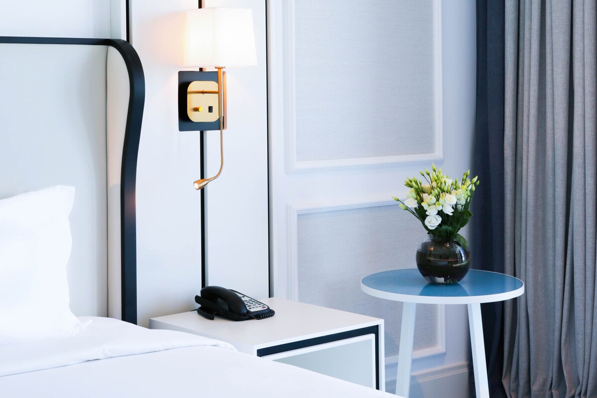A hotel room with a bed, bedside table with a lamp, a phone, and a small table with a vase of flowers.