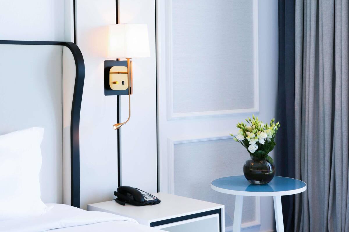A hotel room with a bed, a bedside phone, a lamp, a vase with flowers, and a small table.