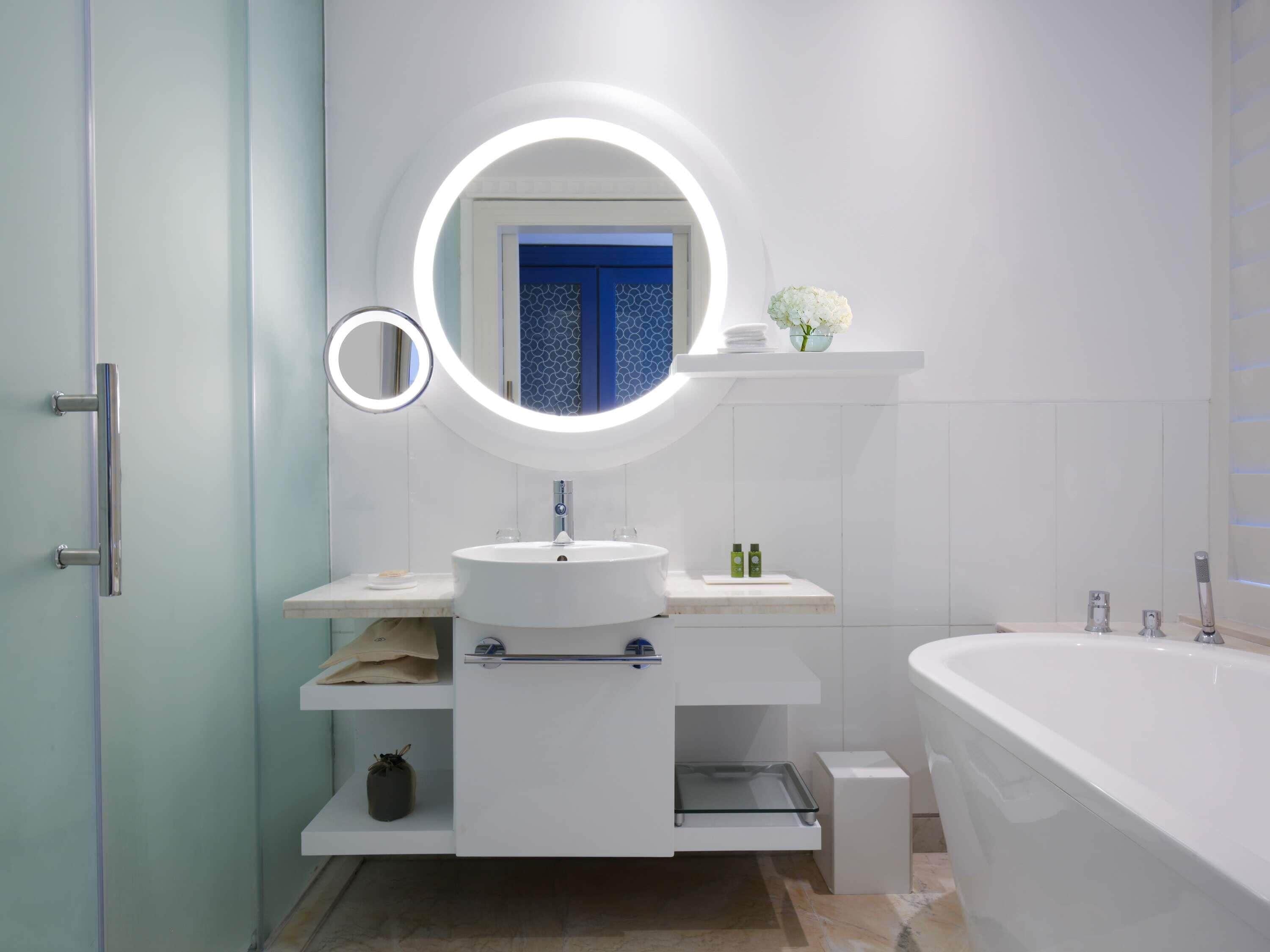 Ein modernes Badezimmer mit einem runden Spiegel, einem Waschbecken, einer Badewanne und einem Ablagefach mit Pflegeprodukten.