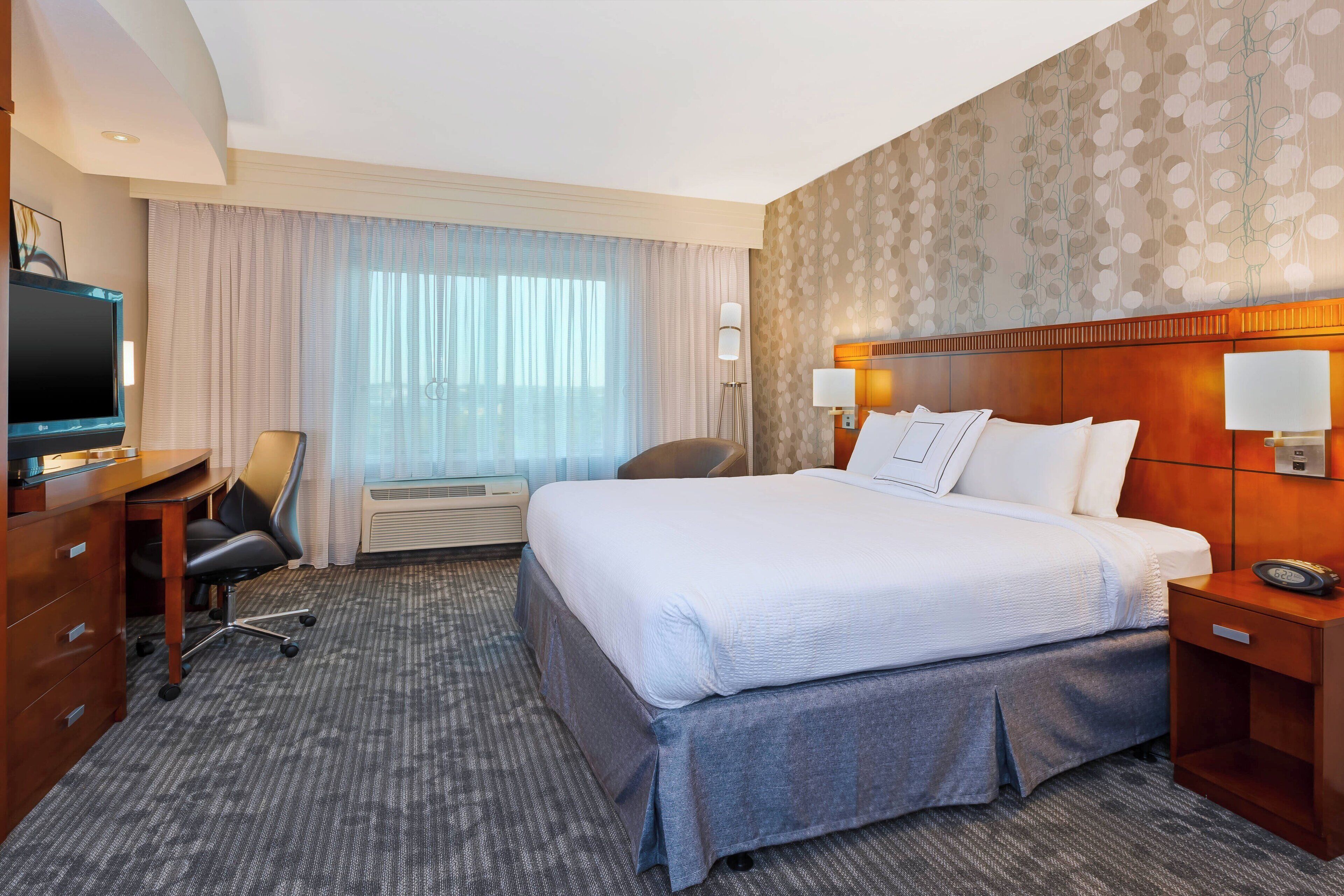 Une chambre d’hôtel avec un grand lit, un bureau avec une chaise, une télévision et une fenêtre avec des rideaux.