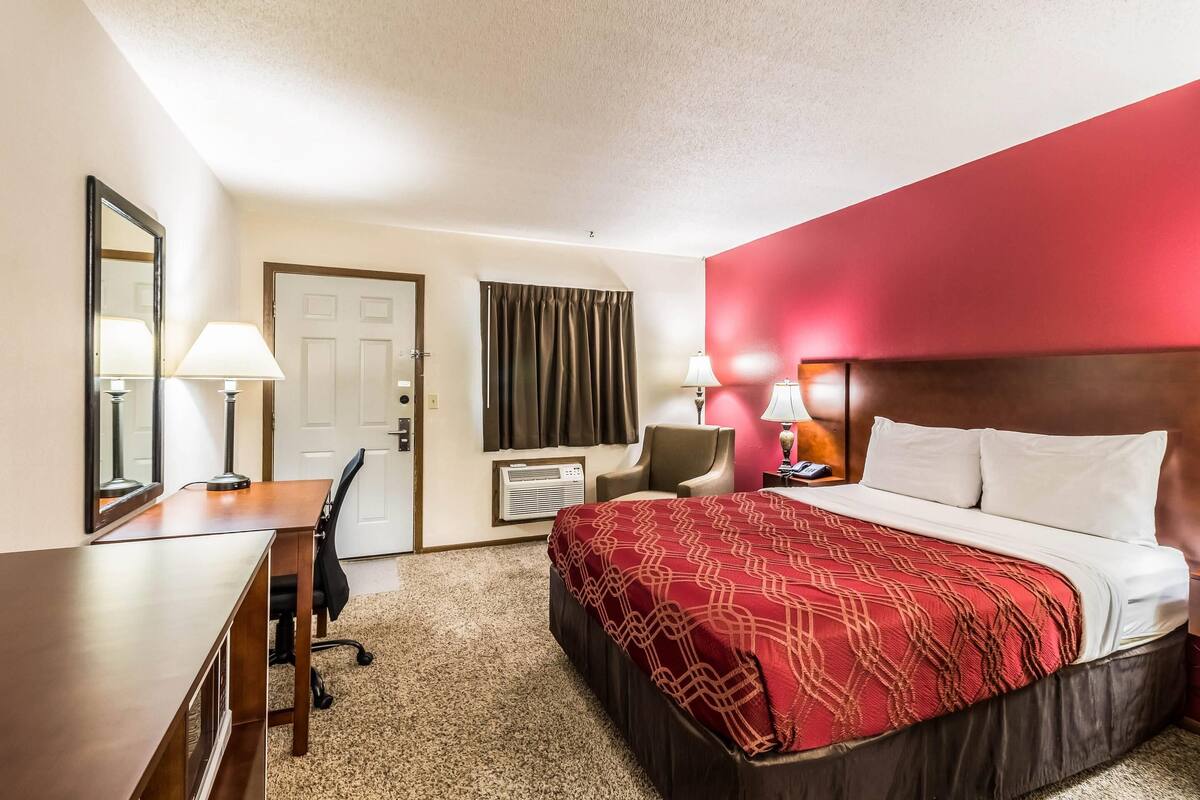 A hotel room with a bed, desk, chair, and a red accent wall.
