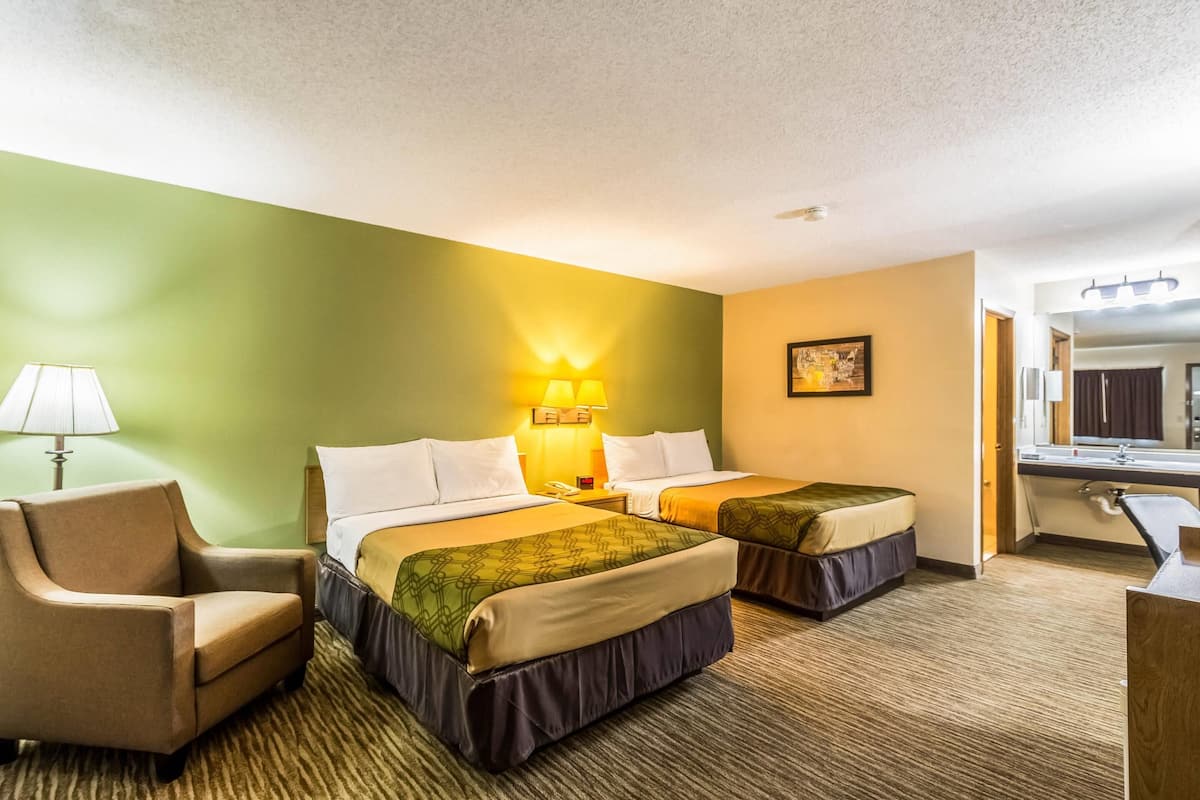 A hotel room with two beds, a chair, a lamp, a painting, and a bathroom visible through an open door.