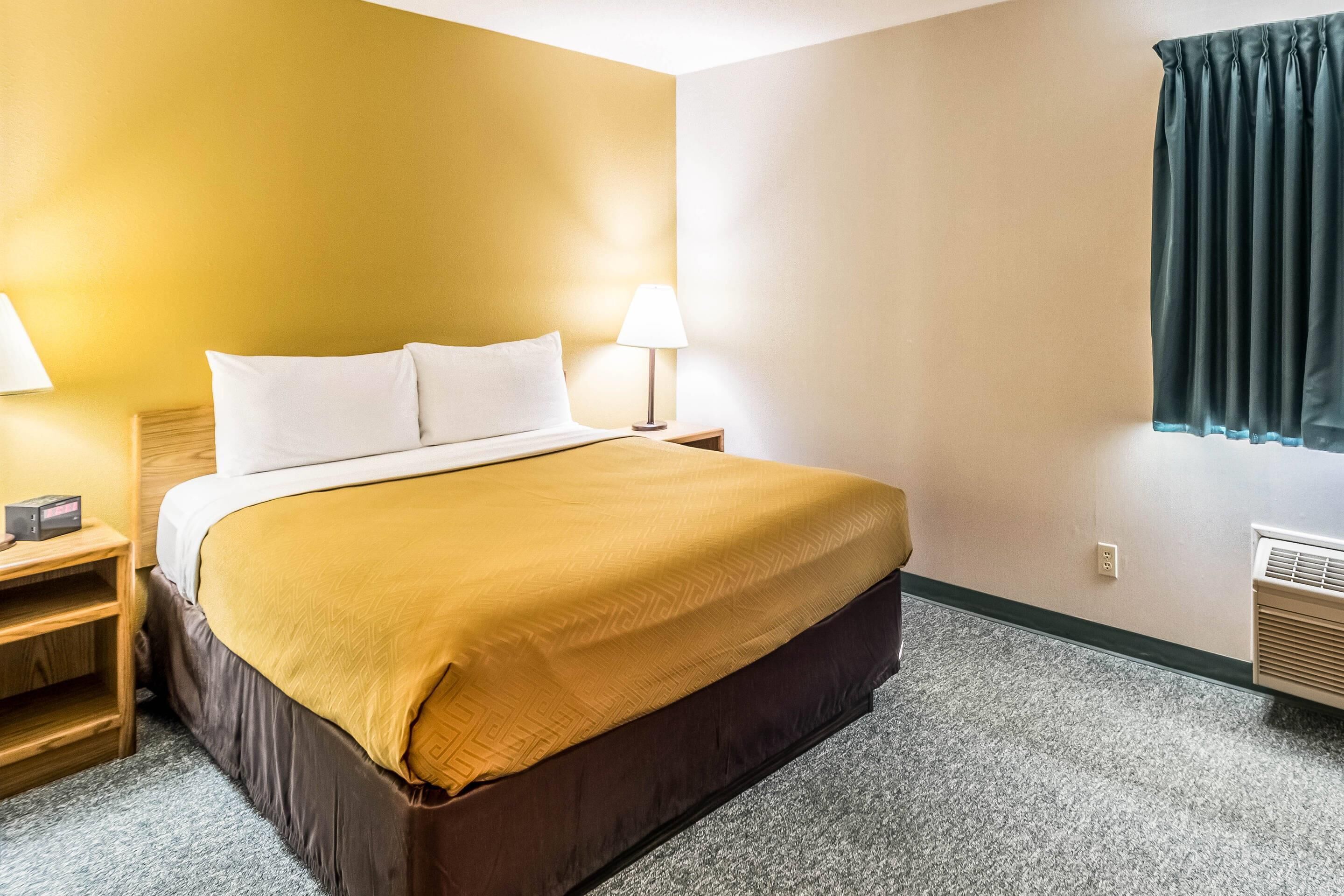 A hotel room with a bed, two bedside tables, a lamp, a window with curtains, and an air conditioning unit.