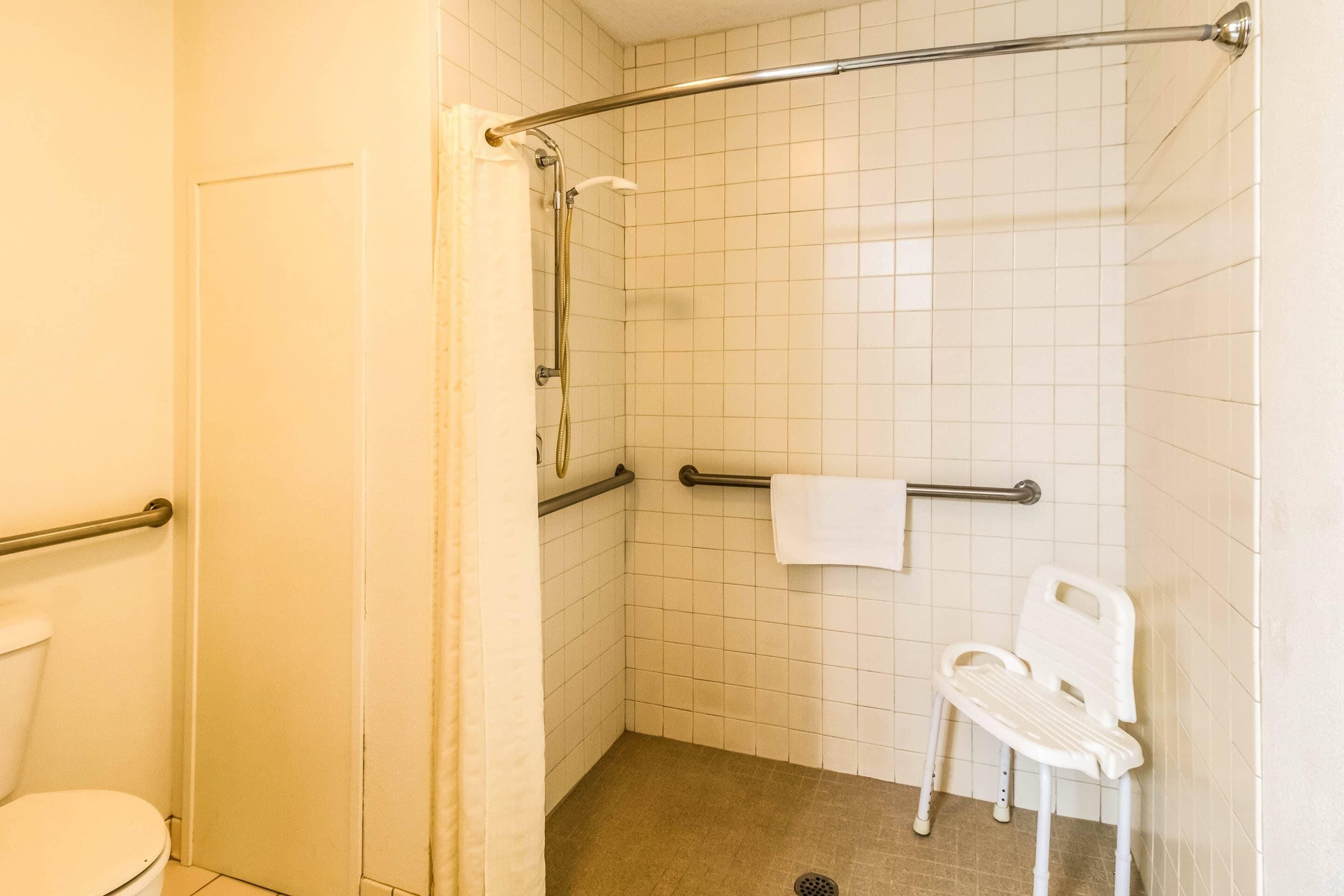 A small, tiled bathroom with a shower, a toilet, a grab bar, and a white plastic chair.