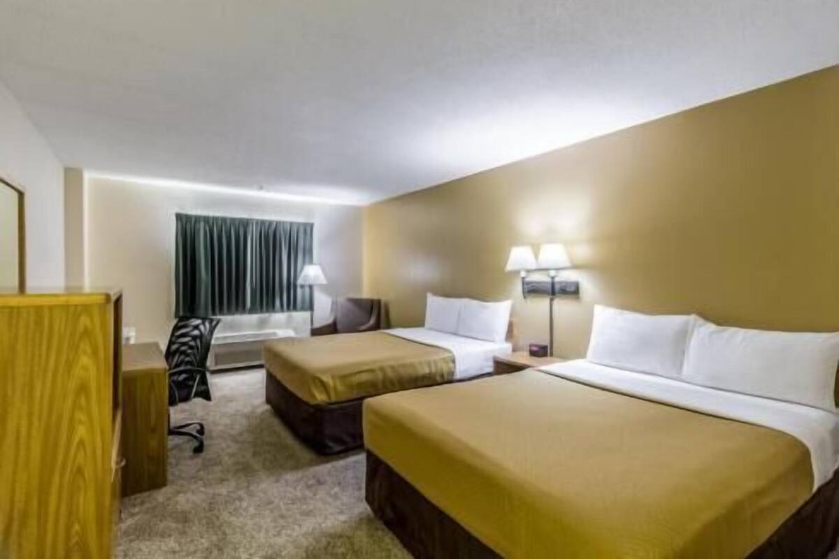 A hotel room with two beds, a desk, a chair, a lamp, and a window with curtains.