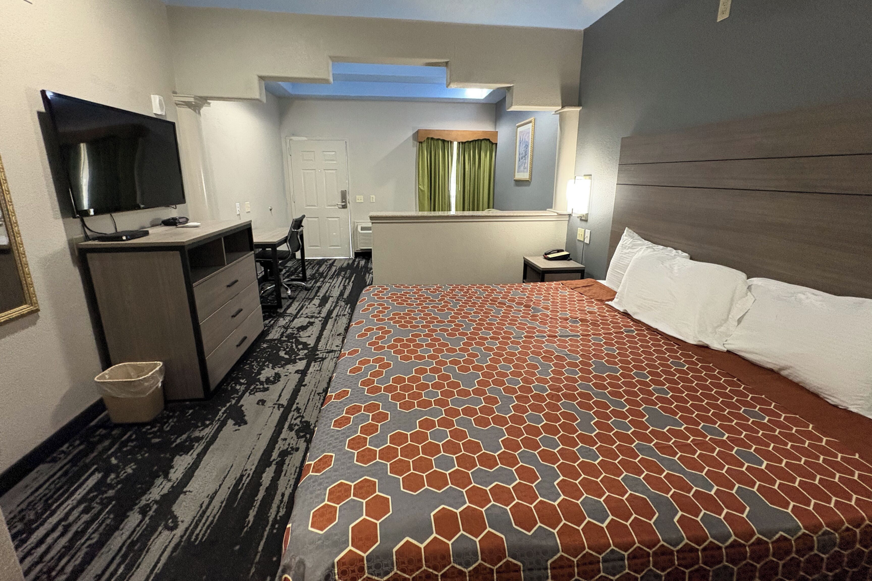 A hotel room with a large bed, a desk with a TV, a chair, and a small table with a lamp.